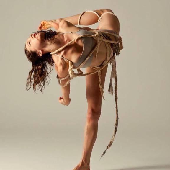 Dancer arching back and kicking leg to meet her head wearing a nude costume with strings of fabric hanging off
